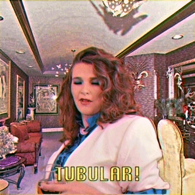 Video gif. A woman wearing a white suit and blue eyeshadow is carrying a drink in one hand and giving us a thumbs up in the other. She winks and the text below reads, "Tubular!"