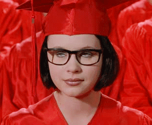 Movie gif. Thora Birch as Enid in Ghost World. She has glasses on and is wearing a red graduation cap and gown and she rolls her eyes while waiting with the other graduates.