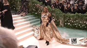 Met Gala 2024 gif. Slow zoom out on Serena Williams, dressed in gold, posing for the cameras with dignified glamor.