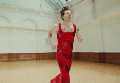 Music video gif. Harry Styles in his video for As It Was wears a red sequined jumpsuit as he jogs wearily in a cavernous ballroom.
