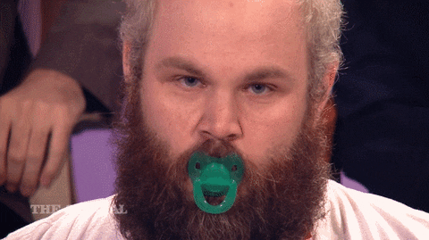 Video gif. A bearded man with a baby's pacifier in his mouth stares at us with a flat expression on his face while he sucks his binky.