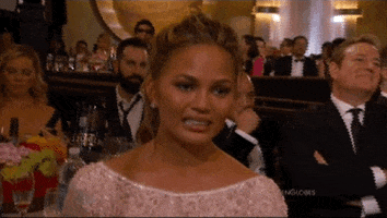 Celebrity gif. Chrissy Teigen at the Golden Globes sits in the audience. She grits her teeth and blankly stares as she grimaces at something awkward.  