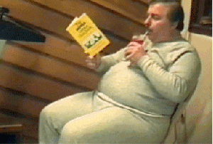 Video gif. A heavyset man in gray sweats relaxes in a wooden chair, reading a book while sipping a drink through a straw. We tilt down to see that his legs are "running" on a treadmill.
