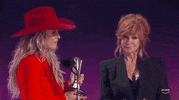 ACM Awards gif. Country artists Lainey Wilson speaks into mic, accepting the thropy she was awarded as Reba McIntre stands nearby with a smile on her face listening attentively 