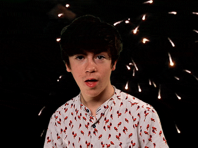 Celebrity gif. A young Declan McKenna wears red, white, and blue as he gives two thumbs up and a wink while fireworks erupt in the background.