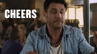 Movie gif. Jaason Simmons as Baz Hogan in Sharknado sits at a bar in a busy restaurant. He holds his beer up and pretends to clink it towards someone. He nods his head with a look of “screw it” on his face and says, “cheers.” 