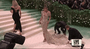 Met Gala 2024 gif. Jennifer Lopez wearing a custom Schiaparelli Haute Couture gown, golden and fully beaded with a sky-high plunging neckline akin to butterfly wings, adjusts herself, getting comfortable and checking in with her cleavage to confirm her breasts are still in place before posing for the cameras.