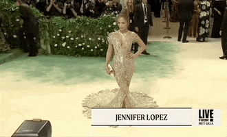 Met Gala 2024 gif. JLo poses with one hand on her hip showing the full view of her Schiaparelli nearly see-through silver foil gown with a concentrated detailing 