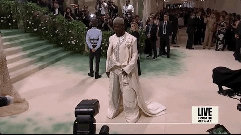 Met Gala 2024 gif. Lil Nas X poses on the red carpet striking a contemplative pose with one hand held to his forehead. He's wearing a monochrome ivory-colored Luar suit with a full trench coat that falls and drapes slightly along the floor. There is a curved geometric line of buttons down the front of his tank top that is decorated in thousands of Swarovski crystals. 