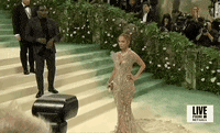 Met Gala 2024 gif. We zoom in on Jennifer Lopez posing with one hand on her hip and a stoic yet confident expression. She's wearing a nearly-see-through Schiaparelli sleeveless gown with a plunging v-neckline that culminates in a pattern that flares out from her shoulders like butterfly wings. She's wearing a prominent diamond necklace, also resembling feathers. 