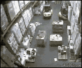 Video gif. Camera footage of a warehouse. An employee is operating a forklift and accidentally swings into a shelf, causing the entire warehouse's stock to fall over with an avalanche of boxes pouring down on them.