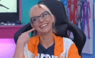 Celebrity gif. Emma Fyffe on Hyper RPG sits in her gamer chair with a cheeky smile on her face. She points to her cheeks and then cutely makes moves her fists up and down while looking around. 