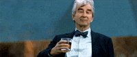Celebrity gif. Sam Waterston in a suit and bowtie slowly holds up a glass of alcohol with a smug expression on his face. The wind rustles through his silver hair. 