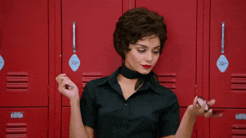 Movie gif. Vanessa Hudgens as Rizzo in Grease Live purses her lips, leaning against lockers, and looking up at herself in a compact mirror.