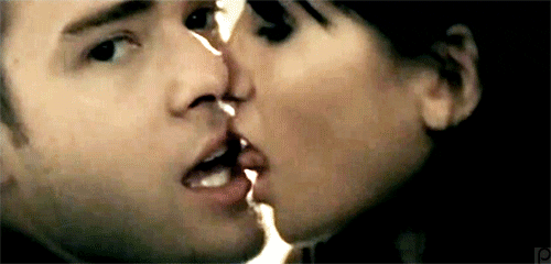 Music video gif. From Cry Me a River, Justin Timberlake looks and smiles at us while a dark-haired woman licks his face sensually.
