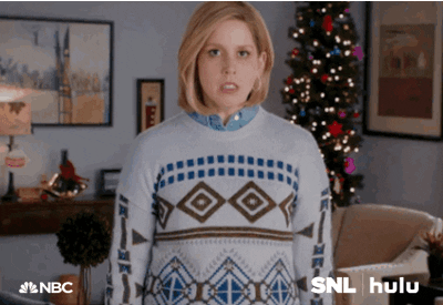 SNL gif. Vanessa Bayer stands in a living room decorated for Christmas looking straight ahead, breathing heavily. She grits her teeth as the shot cuts closer into her face. Suddenly flames erupt around her and in her eyes expressing rage.