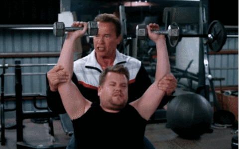 Late Night Show gif. James Cordon holds two normal sized exercise weights over his head as Arnold Schwarzenegger spots him, holding his arms up. James Corden drops the weights and pants exaggeratedly like he’s overly exhausted by the pretty small weights.