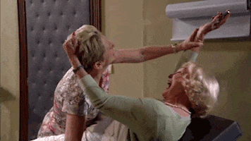TV gif. Character from Thuis with blonde hair who appears to be laying on a medical exam table laughs hysterically as she yanks another woman by the hair, possibly trying to keep her away from the object she is holding in one hand. A man enters the room and in a panic tries to stop the commotion.