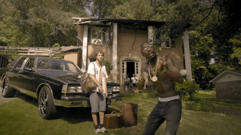 Music video gif. Rich Homie Quan in the Save that Money Music video moves closer to us, putting his hands in a prayer gesture as he begs at us. Behind him is Lil Dicky, leaning on the hood of an older car with luggage around him. In the background is a house with a fallen in roof and men sitting on the stoop.