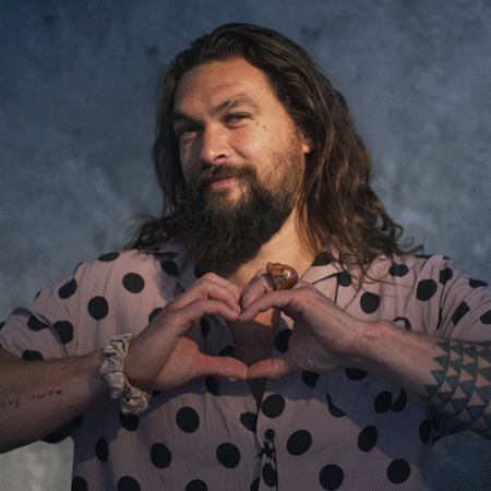 Celebrity gif. Jason Momoa looks at us with a warm smile as he forms his hands into a heart shape. He then places his hands under his chin before going back to forming the heart.