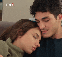 TV gif. Enes Koçak and Eylül Tumbar as Serkan and Ali̇ze in Kendi Düşen Ağlamaz. They're laying on a couch together with their eyes closed. Alize's head rests on Serkan's chest, and Serkan affectionately rubs his cheek against the top of Alize's head as she sleeps peacefully. 