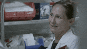 TV gif. Laurie Metcalf as Jenna on Getting On, standing near a shelf holding medical supplies, with a straight face that morphs into a strained smile as she says, "great!" which appears as text.