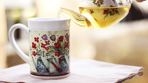 Video gif. A glass teapot pours a gold liquid into a floral tea cup. 