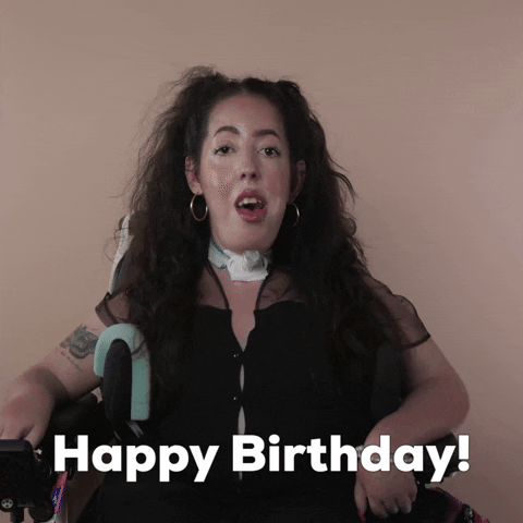 Reaction gif. A Disabled white woman with muscular dystrophy with wavy brown half up half down with two pigtails on top, seated in her motorized wheelchair, bright-eyed, says "Happy birthday!"