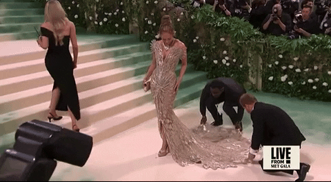 Met Gala 2024 gif. Jennifer Lopez wearing a custom Schiaparelli Haute Couture gown, golden and fully beaded with a sky-high plunging neckline akin to butterfly wings, adjusts herself, getting comfortable and checking in with her cleavage to confirm her breasts are still in place before posing for the cameras.
