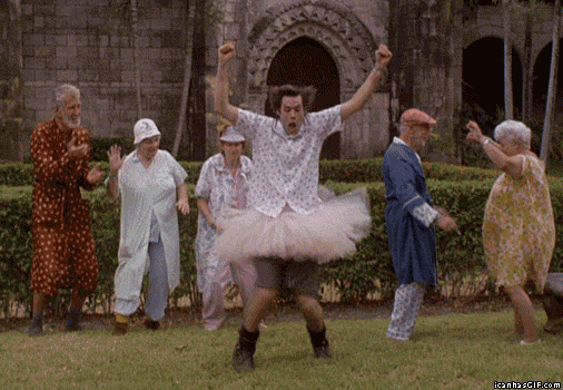 Movie gif. Jim Carrey as Ace in Ace Ventura: Pet Detective wears a tutu, shorts, boots and a button down shirt. His hair is wild, and he holds his hands up in the air as he dances in the yard of an asylum. 