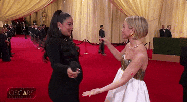 Oscars 2024 GIF. Pre-show hosts Vanessa Hudgens and Julianne Hough hug each other on the red carpet. Hudgens wears a black dress and holds a microphone in her hand while Hough wears a gold and white dress.