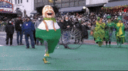 Video gif. Two armless munchkins in costume from Wicked jot through the Macy's Thanksgiving parade.