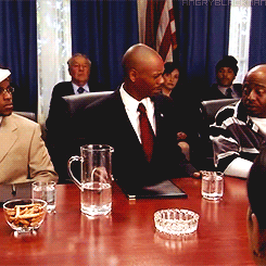 TV gif. Dave Chappelle as Black Bush at a conference desk shakes his head, then knocks over a pitcher of water and jumps up, causing chaos.