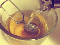 Video gif. Into a glass cup, hot water pours over a stainless steel loose tea infuser.