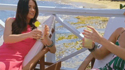 Reality TV gif. Two women from the show Coupled lounge on beach chaises and clink glasses.