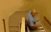 Video gif. An old man rides a chair lift assist down the stairs.