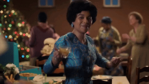 TV gif. Leonie Elliott as Lucille in Call the Midwife lifts a glass toward us and says, "Cheers!" as she bounces away while women dance by a Christmas tree in the background.