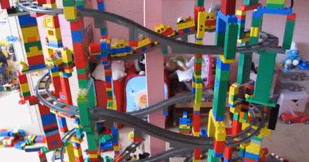 Video gif. Lego train moves on an elaborate, multi-teared built train track and the tower comes crashing down.