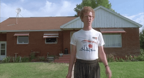 Movie gif. Jon Heder as Napoleon Dynamite stands in a yard in front of a house and raises his hand in an awkward hello.
