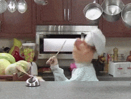 Video gif. Puppet chef in a white apron and a pink bowtie stands behind a kitchen counter. He plays the drums with some watermelons and mixing spoons and he's dancing happily.