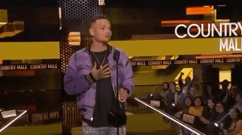 Celebrity gif. Kane Brown at the 2018 American Music Awards steps back from the mic and taps his chest, holding an award with his other hand.