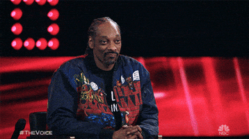 Reality TV gif. Snoop Dogg on The Voice has a smirk on his face and he holds two thumbs up. 