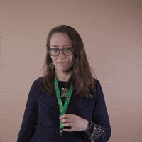 Reaction gif. A Disabled Latina woman with brown wavy hair and glasses shows off her green lanyard, presenting it with an ok sign and a knowing smile.