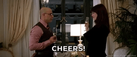 Movie gif. Stanley Tucci as Nigel and Anne Hathaway as Andrea in Devil Wears Prada. They stand in front of each other in a hotel room and clink glasses in success as they say, "Cheers."