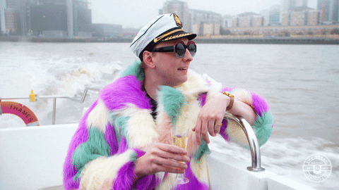 Video gif. Person wearing a sailor hat and a multicolor fur coat holds a champagne as they ride on a boat. He waves at someone.