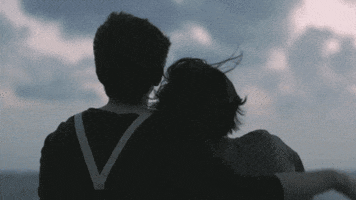 Video gif. Two people sit looking at a cloudy sky. One of the people leans against the other, with their head on the other’s shoulder. The one sitting up straight places their arm gently around the other.