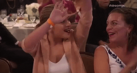 Video gif. At the Streamy Awards a young woman looks over her shoulder and raises her hands in a wave as people around her clap. 