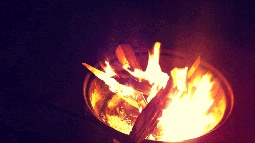 Video gif. Bright flames flicker in a firepit and engulf pieces of overlapping wood.