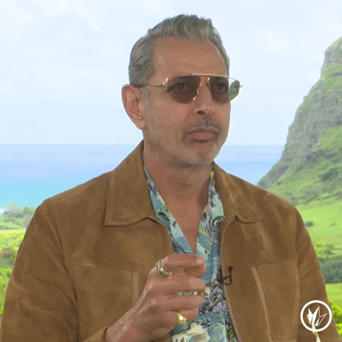 Celebrity gif. Jeff Goldblum in a suede jacket and a tropical-print button-down shirt, standing in front of a cliff and body of water, rolls his eyes and opens his hand.