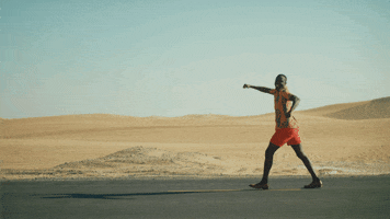 Video gif. A man jauntily walks along a paved road in an empty vast desert. 